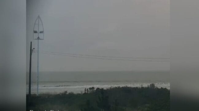 Fotografia em plano geral sob iluminação fraca e tempo fechado em uma praia. O céu e a linha do horizonte sobre o mar estão cinzentos e cobertos por uma névoa densa de tempestade. À esquerda, destaca-se um poste alto com uma estrutura metálica em formato de vela ou barco no topo e fios da rede elétrica cruzando a imagem. Ao fundo, veem-se pequenas silhuetas de pessoas caminhando na faixa de areia próxima às ondas do mar, enquanto a vegetação de restinga ocupa o primeiro plano inferior