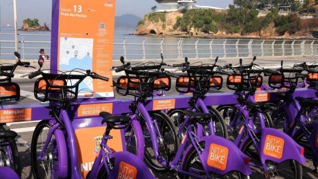 Fotografia diurna em plano médio de uma estação de compartilhamento de bicicletas "NitBike" em Niterói. Diversas bicicletas de cor roxa vibrante, com a identificação "NIT BIKE" e "NITERÓI DE BICICLETA", estão estacionadas em torno de um totem laranja alto. O totem exibe "Estação 13" e "Praia das Flechas", com um mapa, instruções e QR code. Ao fundo, destacam-se a Baía de Guanabara com águas calmas, formações rochosas com vegetação e o Museu de Arte Contemporânea (MAC) de Niterói no topo do morro. O calçadão da orla e uma grade de proteção completam a cena sob céu limpo