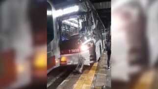 Fotografia na vertical capturada no interior de um terminal de ônibus à noite, mostrando a frente bastante danificada de um ônibus municipal de cor branca e detalhes vermelhos (prefixo 7 3858) após colidir contra a traseira de outro veículo na plataforma. O para-brisa do coletivo está totalmente trincado e estilhaçado, o para-choque dianteiro amassado e o farol esquerdo pendurado próximo ao piso tátil amarelo da plataforma de embarque. À direita, vê-se parte do corpo de um pedestre caminhando pelo local.