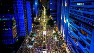 Fotografia aérea noturna em plano aberto capturada do alto de um edifício, mostrando a Avenida Paulista completamente lotada por uma multidão de pessoas. Os arranha-céus que ladeiam a avenida estão iluminados com luzes azuis, enquanto o corredor central da via exibe iluminação amarelada intensa, estruturas de palcos, tendas operacionais e holofotes ao longo da pista