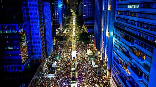 Fotografia aérea noturna em plano aberto capturada do alto de um edifício, mostrando a Avenida Paulista completamente lotada por uma multidão de pessoas. Os arranha-céus que ladeiam a avenida estão iluminados com luzes azuis, enquanto o corredor central da via exibe iluminação amarelada intensa, estruturas de palcos, tendas operacionais e holofotes ao longo da pista