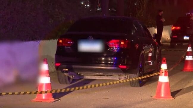 Fotografia noturna em plano médio de uma cena de crime isolada pela polícia em uma via pública. No centro da imagem, observa-se a traseira de um automóvel sedã preto (modelo Volkswagen Jetta) estacionado próximo a um muro, com a placa desfocada. O local está bloqueado por uma fita zebrada amarela e preta e cones de sinalização laranjas com faixas refletivas brancas. Do lado esquerdo, uma grande área da imagem apresenta borrão para preservar conteúdo sensível no solo. Ao fundo, policiais ou peritos em trajes escuros permanecem ao lado de outro veículo