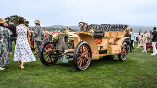 Fotografia em plano três-quartos dianteiro de um automóvel clássico de luxo do início do século 20, modelo American Mercedes de 1905, exposto sobre o gramado de um evento ao ar livre à beira-mar. O veículo tem carroceria bege, assentos de couro preto em estilo capitonê no formato Double Phaeton, grandes faróis e acabamentos em latão dourado polido na grade e na buzina, além de rodas de madeira com raios pintados em vermelho. Ao redor, pessoas com trajes casuais e elegantes caminham pelo gramado sob céu encoberto