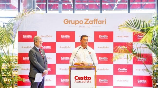 Fotografia em plano médio do evento de inauguração da primeira loja da rede Cestto Atacadista no estado de São Paulo. No centro da imagem, um homem vestindo camisa social branca discursa em um púlpito de madeira que exibe o logotipo do "Cestto Atacadista". À esquerda, um executivo de terno cinza, gravata vermelha e óculos acompanha a apresentação de pé. Ao fundo, destaca-se um grande painel institucional com padrão xadrez em vermelho e branco estampando as marcas "Grupo Zaffari" e "Cestto Atacadista". Folhagens tropicais de palmeiras em vasos emolduram as laterais do palco sob iluminação diurna