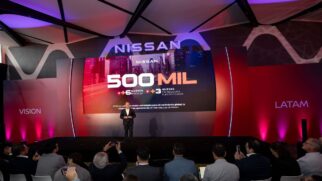 Fotografia em plano geral do palco de um evento corporativo da montadora Nissan para a América Latina. No centro, um executivo discursa diante de um grande telão de LED que exibe em destaque a inscrição "500 MIL", ladeada pelos textos em espanhol "+6 NUEVOS MODELOS" e "+3 NUEVAS TECNOLOGÍAS ELECTRIFICADAS". O painel superior traz o logotipo "NISSAN" e a iluminação do ambiente varia entre tons de roxo, rosa e vermelho, com estruturas laterais indicando "VISION" e "LATAM". Em primeiro plano, a plateia de jornalistas e executivos assiste ao painel e registra a apresentação com smartphones