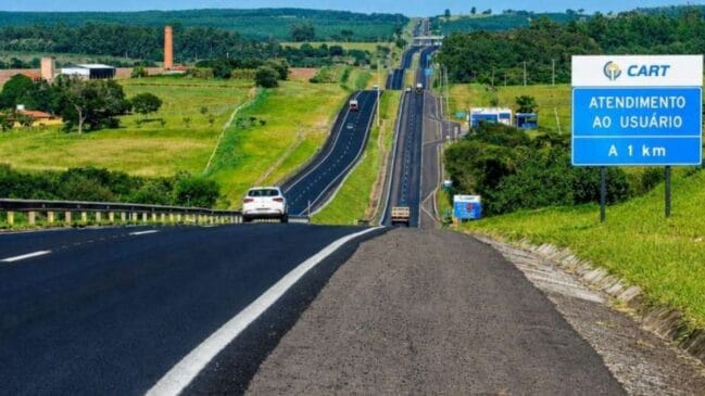 Fotografia diurna em perspectiva de profundidade de uma rodovia duplicada de asfalto escuro (SP-270) que se estende por morros e aclives em uma paisagem rural verde. À direita, em primeiro plano no acostamento, destaca-se uma placa azul da concessionária CART com a inscrição "ATENDIMENTO AO USUÁRIO A 1 km". Na pista, um veículo de passeio branco trafega no mesmo sentido em que a imagem foi capturada, enquanto outros veículos e caminhões aparecem mais ao fundo sob céu ensolarado
