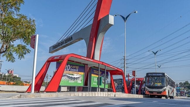 Fotografia em plano geral e iluminação diurna mostrando a estrutura do terminal e parada de ônibus municipal em Taboão da Serra. No centro, destaca-se uma cobertura metálica em tom vermelho brilhante sob uma grande estrutura arquitetônica com o letreiro "TABOÃO DA SERRA" em destaque. No painel do ponto, há cartazes sobre a entrega de novos ônibus para a cidade. À direita, um ônibus urbano de cores branca e alaranjada da frota municipal aguarda o embarque de passageiros. Em primeiro plano, a via exibe sinalização de faixa de pedestres sob céu azul