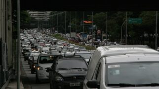 Fotografia em plano longo sob a estrutura de um viaduto, mostrando congestionamento intenso de veículos na Avenida 23 de Maio em São Paulo. A pista está repleta de carros, táxis brancos e furgões alinhados em várias faixas
