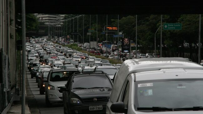 Fotografia em plano longo sob a estrutura de um viaduto, mostrando congestionamento intenso de veículos na Avenida 23 de Maio em São Paulo. A pista está repleta de carros, táxis brancos e furgões alinhados em várias faixas