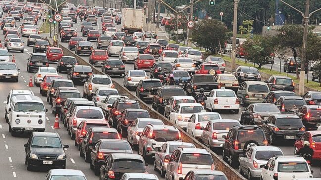 Fotografia em plano geral com lente teleobjetiva mostrando engarrafamento em uma avenida de múltiplas faixas. O tráfego está completamente parado ou em marcha lenta, com carros de passeio, táxis, uma perua Kombi branca à esquerda e motocicletas enfileiradas entre as faixas. No canteiro central arborizado, observam-se placas de sinalização indicando o limite de velocidade de 60 km/h e luzes de freio vermelhas acesas ao longo de toda a extensão da via sob luz diurna