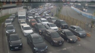 Fotografia de câmera de monitoramento do bolsão de embarque da travessia de balsas entre Santos e Guarujá. Múltiplas filas paralelas de carros de passeio de diversas cores estão paradas aguardando o embarque em uma área asfaltada sob dia nublado