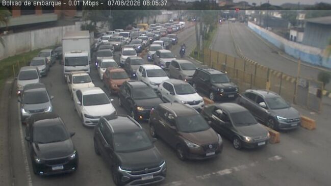 Fotografia de câmera de monitoramento do bolsão de embarque da travessia de balsas entre Santos e Guarujá. Múltiplas filas paralelas de carros de passeio de diversas cores estão paradas aguardando o embarque em uma área asfaltada sob dia nublado
