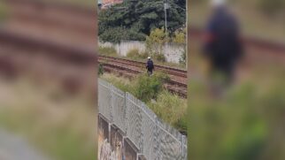 Fotografia em plano aberto e levemente elevado mostrando a faixa de domínio de uma ferrovia. Ao centro, um funcionário vestindo capacete de proteção branco, uniforme escuro com faixa refletiva e mochila caminha sobre o britamento entre os trilhos da linha férrea