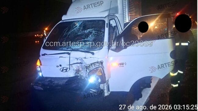 Fotografia noturna em plano frontal de um caminhão utilitário leve de carga (VUC) na cor branca, modelo Hyundai HR com baú, acidentado e parado na pista. O veículo apresenta severe avaria na extremidade dianteira esquerda, com para-choque quebrado, grade frontal amassada, porta do motorista aberta e para-brisa totalmente estilhaçado em teia de aranha. À direita da imagem, um agente de resgate com faixa refletiva no uniforme atua no local. Marcas d'água da Artesp e o registro impresso com a data "27 de ago. de 2026 03:15:25" estão visíveis na imagem