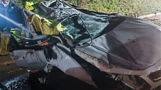 Fotografia noturna em plano médio de um automóvel de passeio na cor branca completamente destruído após capotamento. O veículo está com o teto totalmente esmagado, para-brisa estilhaçado, capô retorcido e a lataria da porta lateral amassada. À esquerda, socorristas do Corpo de Bombeiros em equipamentos de proteção e coletes refletivos amarelos atuam na ocorrência. O asfalto aparece molhado refletindo a iluminação de emergência e, ao fundo, observa-se um barranco com vegetação no acostamento da pista