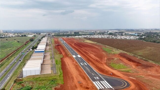 Fotografia aérea diurna mostrando a pista de pouso asfaltada de um aeródromo com a marcação de cabeceira "12" em branco no primeiro plano. A pista é cercada por terra vermelha de obras de expansão, com uma fileira de hangares alinhados à esquerda e uma rodovia movimentada correndo paralelamente ao complexo