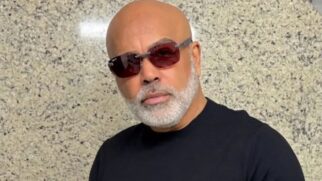 Retrato em plano próximo do cantor Rick, careca e com barba grisalha aparada, a usar óculos de sol com lentes avermelhadas e t-shirt preta diante de uma parede de granito