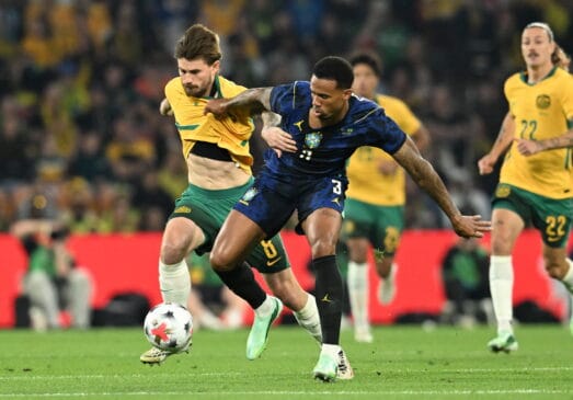 Fotografia de ação de uma partida de futebol entre as seleções do Brasil e da Austrália, mostrando um jogador brasileiro com o equipamento azul a disputar a bola com um adversário australiano de amarelo no relvado do estádio