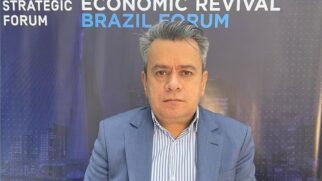 Fotografia em plano médio de palestrante homem com cabelos grisalhos penteados para trás, vestindo paletó azul e camisa social listrada de azul e branco. Ao fundo, painel institucional escuro exibe em destaque os títulos em inglês "World Strategic Forum", "Mastering the Economic Revival" e "Brazil Forum"