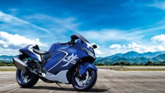 Fotografia diurna em plano geral de uma motocicleta superesportiva Suzuki Hayabusa azul e branca com ideograma japonês na carenagem, estacionada na pista de um aeródromo com montanhas e céu ensolarado ao fundo