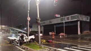 Fotografia noturna de um grave acidente de trânsito num cruzamento urbano, mostrando uma carrinha branca com a parte frontal totalmente destruída e destroços espalhados pela via junto a uma passagem de peões, sob a iluminação de semáforos e postes de luz