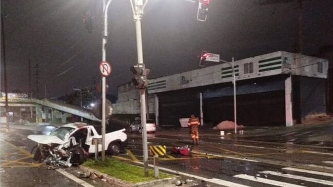 Fotografia noturna de um grave acidente de trânsito num cruzamento urbano, mostrando uma carrinha branca com a parte frontal totalmente destruída e destroços espalhados pela via junto a uma passagem de peões, sob a iluminação de semáforos e postes de luz