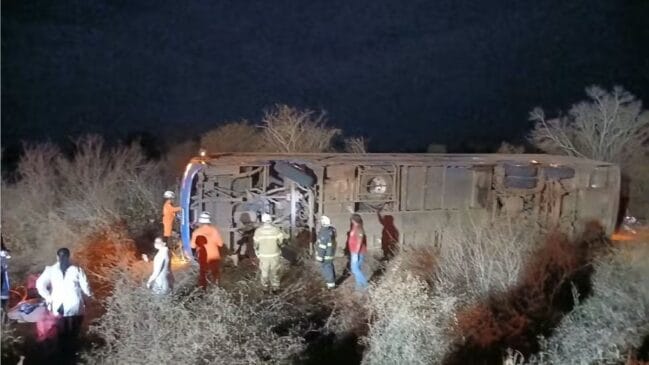 Fotografia noturna de um autocarro de passageiros tombado de lado fora da estrada numa área de mato na Bahia, com equipas de resgate e bombeiros a prestar socorro no local
