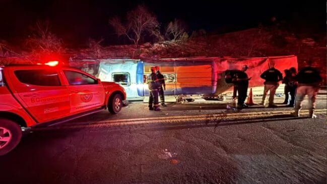 Fotografia noturna de um autocarro tombado de lado numa rodovia no Espírito Santo, com uma viatura de socorro dos bombeiros com luzes de emergência vermelhas acesas e equipas de resgate no local