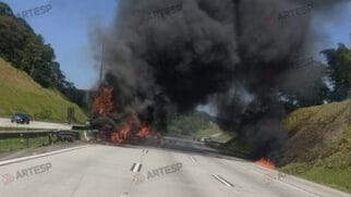 Sugestão: Fotografia de um veículo em chamas com uma densa e elevada coluna de fumo negro a subir ao céu na faixa de rodagem do Rodoanel Mário Covas, com marcas de água da Artesp sobrepostas na imagem