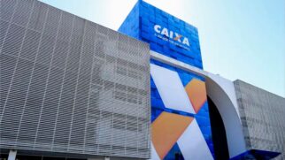 Fotografia diurna em ângulo inferior (de baixo para cima) mostrando a fachada de um prédio moderno da Caixa Econômica Federal. A estrutura possui painel frontal azul com o logotipo "CAIXA" impresso em branco, um grande símbolo do banco estilizado em "X" nas cores laranja e branca, estrutura com brises metálicos e janelas de vidro no térreo sob céu limpo.
