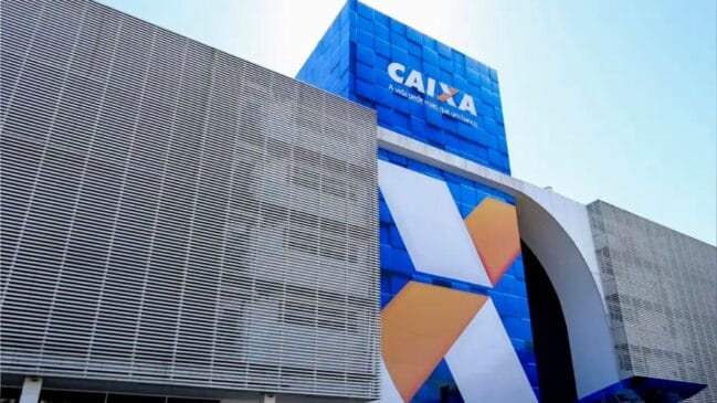 Fotografia diurna em ângulo inferior (de baixo para cima) mostrando a fachada de um prédio moderno da Caixa Econômica Federal. A estrutura possui painel frontal azul com o logotipo "CAIXA" impresso em branco, um grande símbolo do banco estilizado em "X" nas cores laranja e branca, estrutura com brises metálicos e janelas de vidro no térreo sob céu limpo.
