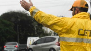 Fotografia em plano médio de um agente de trânsito da CET de São Paulo, visto de costas em ângulo lateral. Ele veste capa de chuva e boné amarelos com a sigla "CET" impressa e está com o braço direito estendido fazendo sinal de parada ou orientação. Ao fundo, desfocado, observam-se carros em circulação numa via pública sob tempo chuvoso