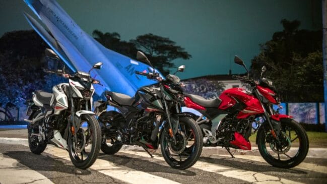 Fotografia de três motocicletas Bajaj Pulsar N150 nas cores branca, preta e vermelha alinhadas lado a lado à noite, posicionadas em frente a uma aeronave monumento da Força Aérea Brasileira iluminada em azul ao fundo