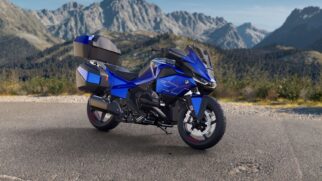 Fotografia diurna de motocicleta touring BMW azul metálico com baús laterais e traseiro instalados, estacionada no acostamento de uma estrada asfaltada com montanhas ao fundo sob céu limpo