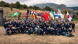 Fotografia de um grupo numeroso de participantes e equipas do BMW Motorrad GS Trophy reunidos ao ar livre num campo relvado com montanhas ao fundo, segurando bandeiras de diversos países, incluindo Brasil, Alemanha, China, Japão e Ucrânia