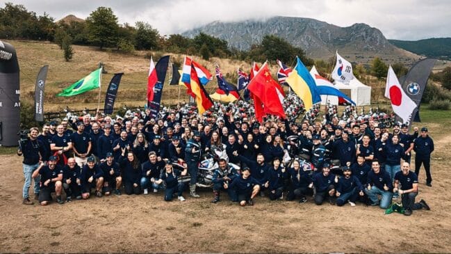 Fotografia de um grupo numeroso de participantes e equipas do BMW Motorrad GS Trophy reunidos ao ar livre num campo relvado com montanhas ao fundo, segurando bandeiras de diversos países, incluindo Brasil, Alemanha, China, Japão e Ucrânia