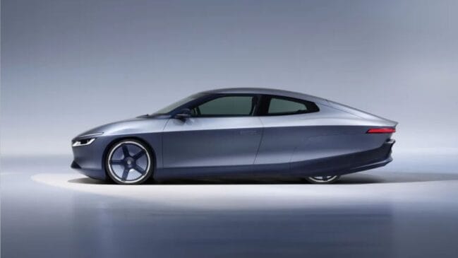 Fotografia de estúdio em perfil lateral de um carro-conceito futurista da Volkswagen na cor cinza-azulado metálico. O veículo possui silhueta aerodinâmica fluida estilo fastback, rodas traseiras encobertas pela carenagem da carroceria, rodas dianteiras com design esportivo e logotipo da marca, maçanetas embutidas e luzes afiladas em LED na dianteira e traseira