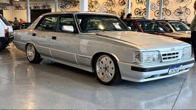 Fotografia de um automóvel clássico Chevrolet Opala Diplomata SE prateado em exposição num salão coberto, com bicicletas vintage penduradas na parede ao fundo e outros veículos antigos em redor