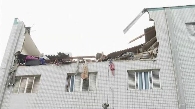 Fotografia em plano fechado da fachada de um prédio residencial de azulejos claros, mostrando o último andar e o telhado parcialmente destruídos após uma explosão. A parede superior cedeu, expondo a estrutura interna do apartamento, entulhos e ferragens sob céu nublado