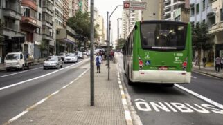 Fotografia diurna em plano médio vista por trás de um ônibus urbano verde do sistema municipal de transporte trafegando por uma faixa exclusiva demarcada no asfalto. Na pista, a sinalização horizontal exibe a palavra "ÔNIBUS" pintada em grandes letras brancas ao lado de uma linha contínua de divisão de faixa. À esquerda, uma calçada/canteiro divisor conta com pedestres caminhando e vias paralelas com tráfego de carros e pequenos caminhões. O entorno é composto por prédios residenciais e comerciais altos sob luz natural diurna