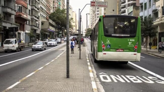 Fotografia diurna em plano médio vista por trás de um ônibus urbano verde do sistema municipal de transporte trafegando por uma faixa exclusiva demarcada no asfalto. Na pista, a sinalização horizontal exibe a palavra "ÔNIBUS" pintada em grandes letras brancas ao lado de uma linha contínua de divisão de faixa. À esquerda, uma calçada/canteiro divisor conta com pedestres caminhando e vias paralelas com tráfego de carros e pequenos caminhões. O entorno é composto por prédios residenciais e comerciais altos sob luz natural diurna