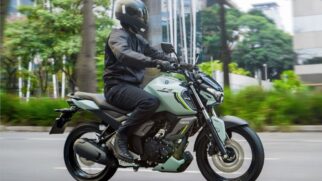 Fotografia em perfil lateral e em movimento de um motociclista usando capacete fechado e vestuário preto pilotando uma motocicleta Yamaha Fazer FZ15 na cor verde-claro fosco. O veículo trafega por uma via urbana asfaltada com arborização e prédios ao fundo