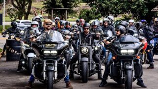 Fotografia frontal de um grupo de motociclistas pilotando motocicletas custom e touring pretas alinhadas em uma via pública, todos usando capacetes e trajes de pilotagem.