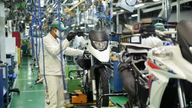 Operário de máscara e luvas a trabalhar na montagem do guiador de uma motocicleta branca na linha de produção de uma fábrica