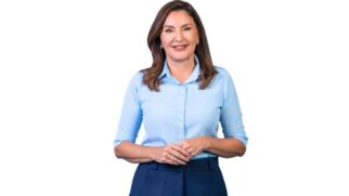Retrato em plano médio da política Hana Ghassan, vice-governadora do Pará, contra um fundo branco neutro. Ela possui cabelos castanhos na altura dos ombros, sorri para a câmera e veste camisa social de manga longa dobrada na cor azul-clara e calça azul-escura, mantendo as mãos cruzadas na altura da cintura