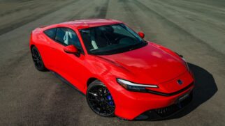 Fotografia em ângulo elevado três quartos dianteiro de um cupê esportivo Honda Prelude na cor vermelha. O veículo exibe faróis afilados em LED, logotipo da Honda na parte frontal, rodas de liga leve pretas com pinças de freio azuis e silhueta aerodinâmica sobre piso de asfalto