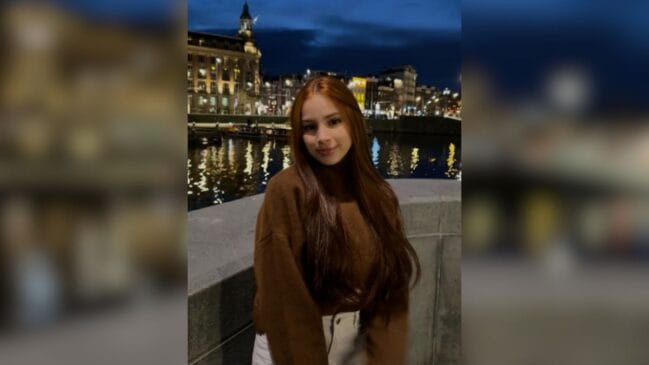 Fotografia em plano médio da influenciadora Duda Alves com longos cabelos castanho-avermelhados e blusa de frio marrom. Ela sorri suavemente posando em uma mureta à noite, com o reflexo de luzes urbanas em um canal de água e edifícios iluminados ao fundo