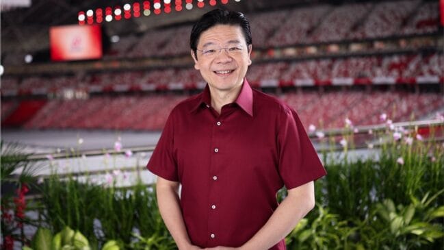 Fotografia em plano médio do primeiro-ministro de Cingapura, Lawrence Wong, vestindo camisa social de mangas curtas na cor vinho e óculos. Ele sorri com as mãos unidas à frente da cintura, posicionado diante de um canteiro com plantas tropicais e com as arquibancadas vermelhas de um estádio ao fundo.