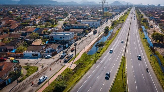 Fotografia aérea diurna em plano panorâmico mostrando a Rodovia Padre Manoel da Nóbrega (SP-055) no litoral paulista. À direita, a rodovia asfaltada exibe tráfego fluido de veículos em múltiplas faixas; à esquerda, observa-se uma via marginal com ciclovia, casas do bairro residencial e morros no horizonte sob céu claro