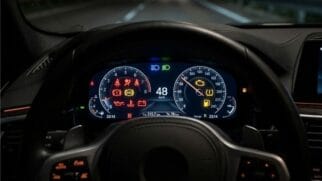 Fotografia em plano próximo do painel de instrumentos digital de um automóvel aceso à noite, exibindo velocímetro, conta-rotações e diversos indicadores luminosos de aviso no mostrador